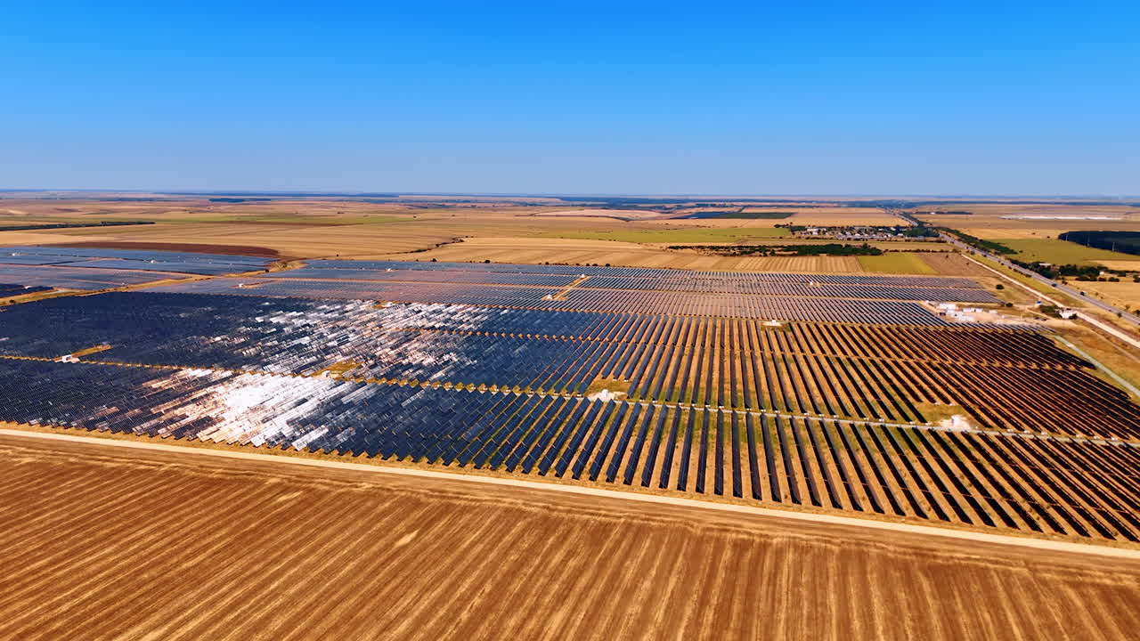 Solar power plant across dry agricultural landscape. Rows of solar panels arranged in large blocks covering open farmland under bright sky