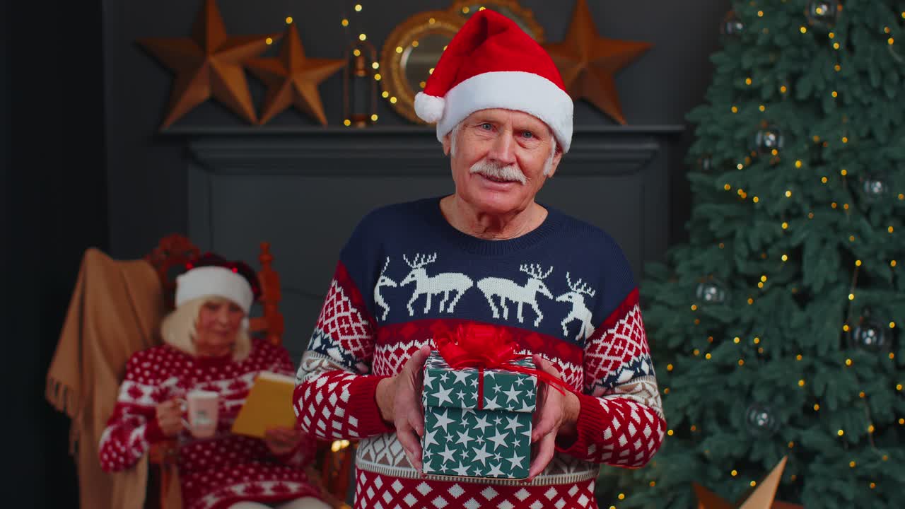 Elderly grandfather in festive sweater presenting christmas gift box smiling looking at camera