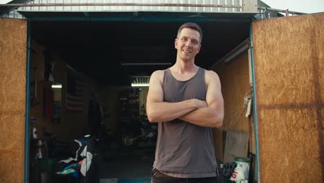 A-happy-male-mechanic-in-a-gray-T-shirt-has-his-hands-folded-on-his-chest,-standing-against-the-backdrop-of-an-open-garage-workshop.-Portrait-of-a-mechanic-near-his-workshop