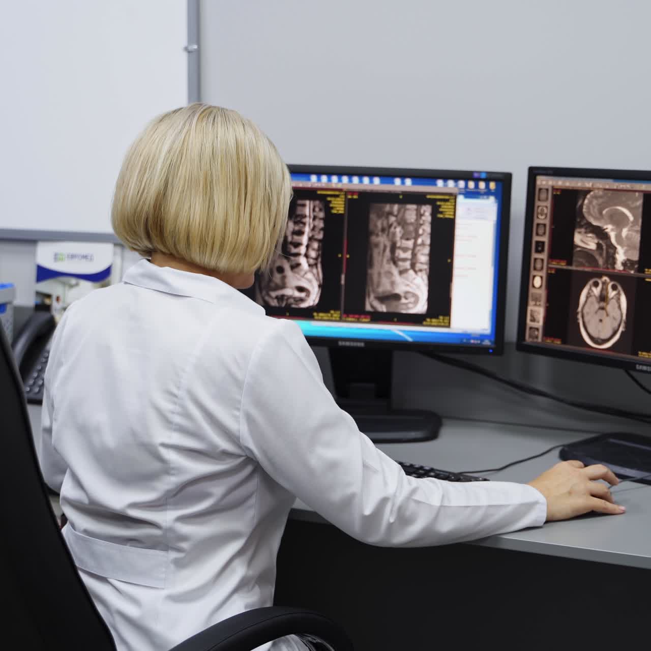 Doctor working in the hospital laboratory for MRI assessment. Female in white robe sits her back to camera looking at scans on the computer