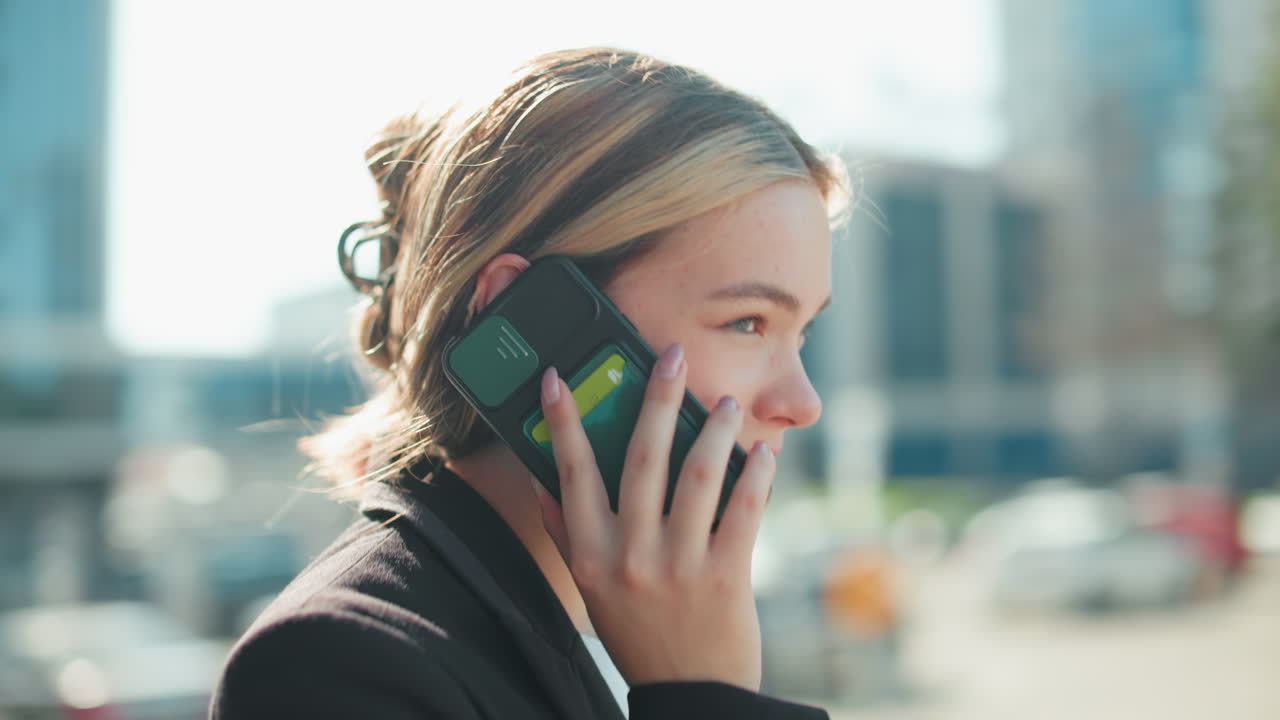 Elegant lady with warm smile conversing over phone with business associate while exiting conference meeting, captured through glass door with urban background reflecting cityscape