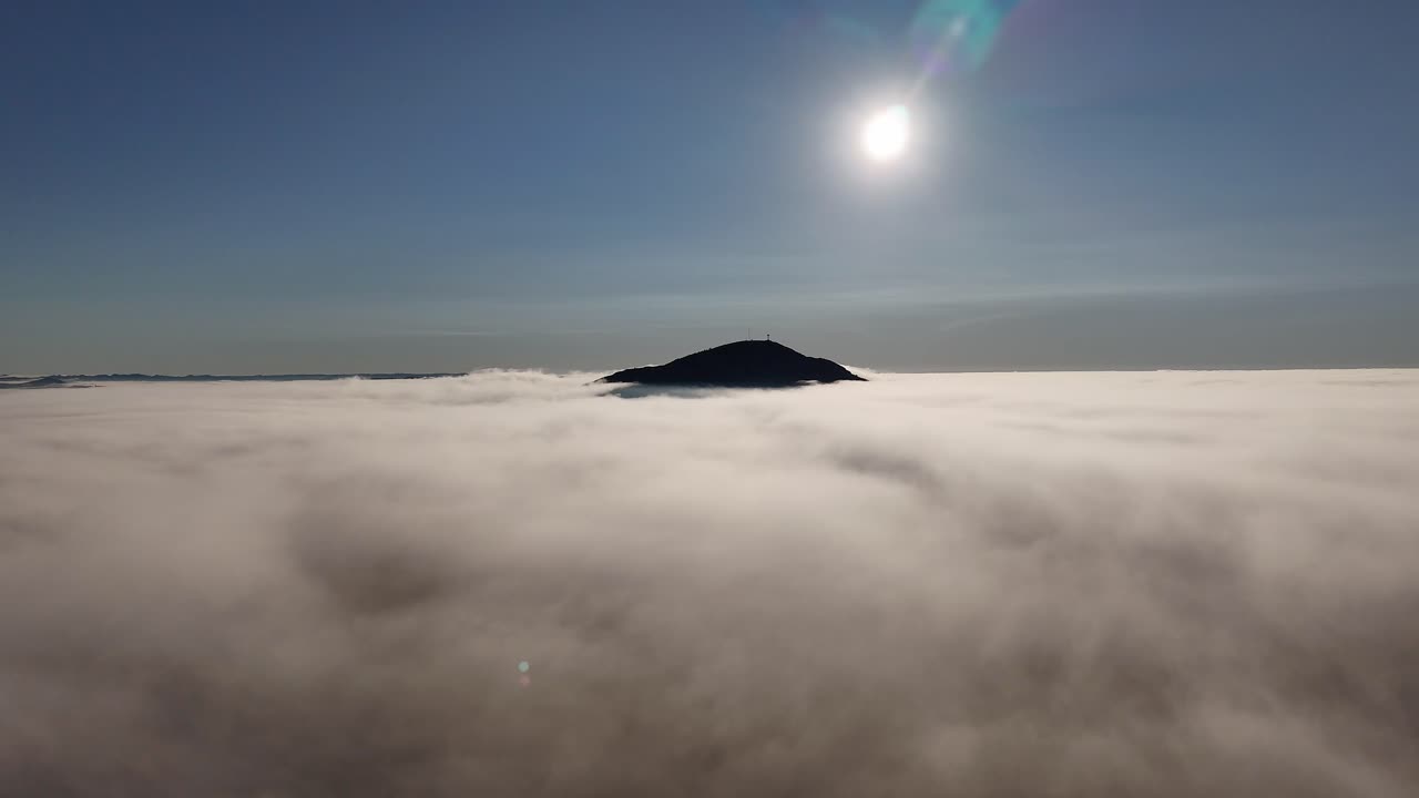 Drone flies above Cerro Pan de Azúcar in Maldonado, Uruguay. A wide open view reveals low clouds and bright sun directly in front, creating a glowing atmospheric landscape