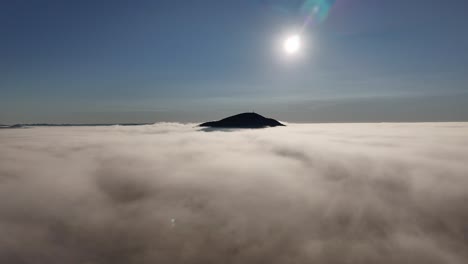drone flies above cerro pan de azúcar in maldonado, uruguay. a wide open view reveals low clouds and bright sun directly in front, creating a glowing atmospheric landscape
