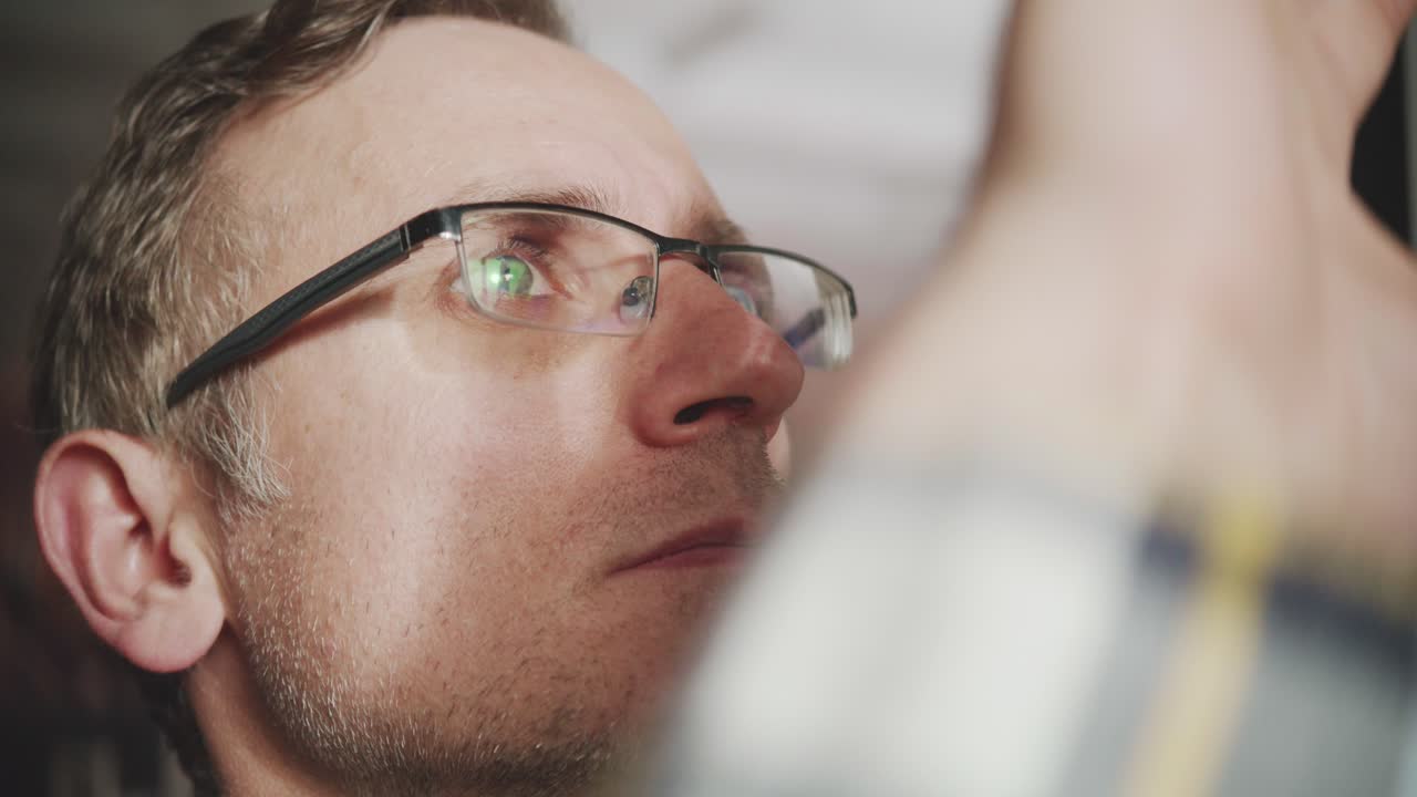An extreme close-up on the face of a man concentrating on his work. The computer monitor and data are reflected in his glasses. A concept of focus, analysis, and technology