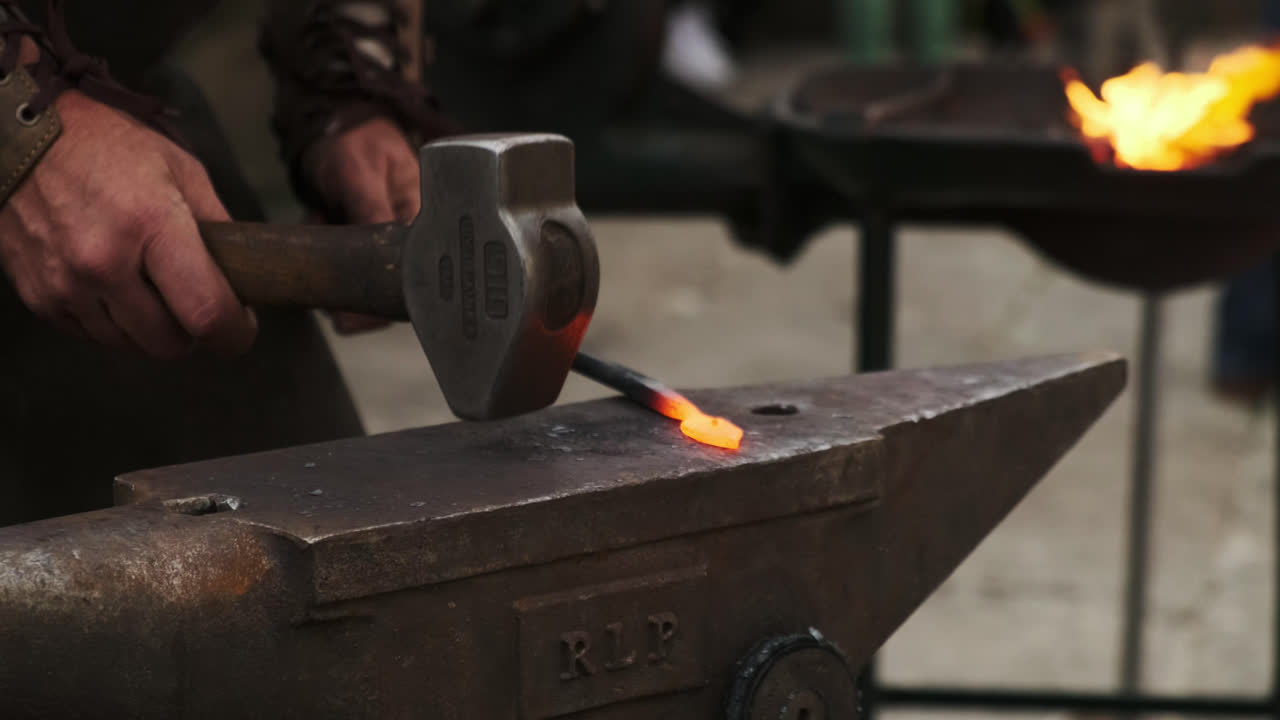 Static close-up of a blacksmith striking red-hot iron on an anvil with a hammer; the coal forge blazes softly out of focus in the background