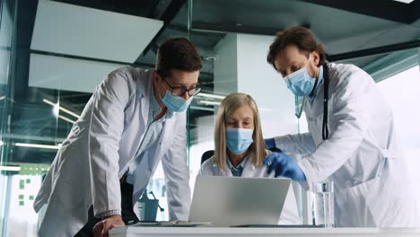 caucasian senior female doctor in medical mask sitting in hospital, typing on laptop and speaking with male colleagues at work