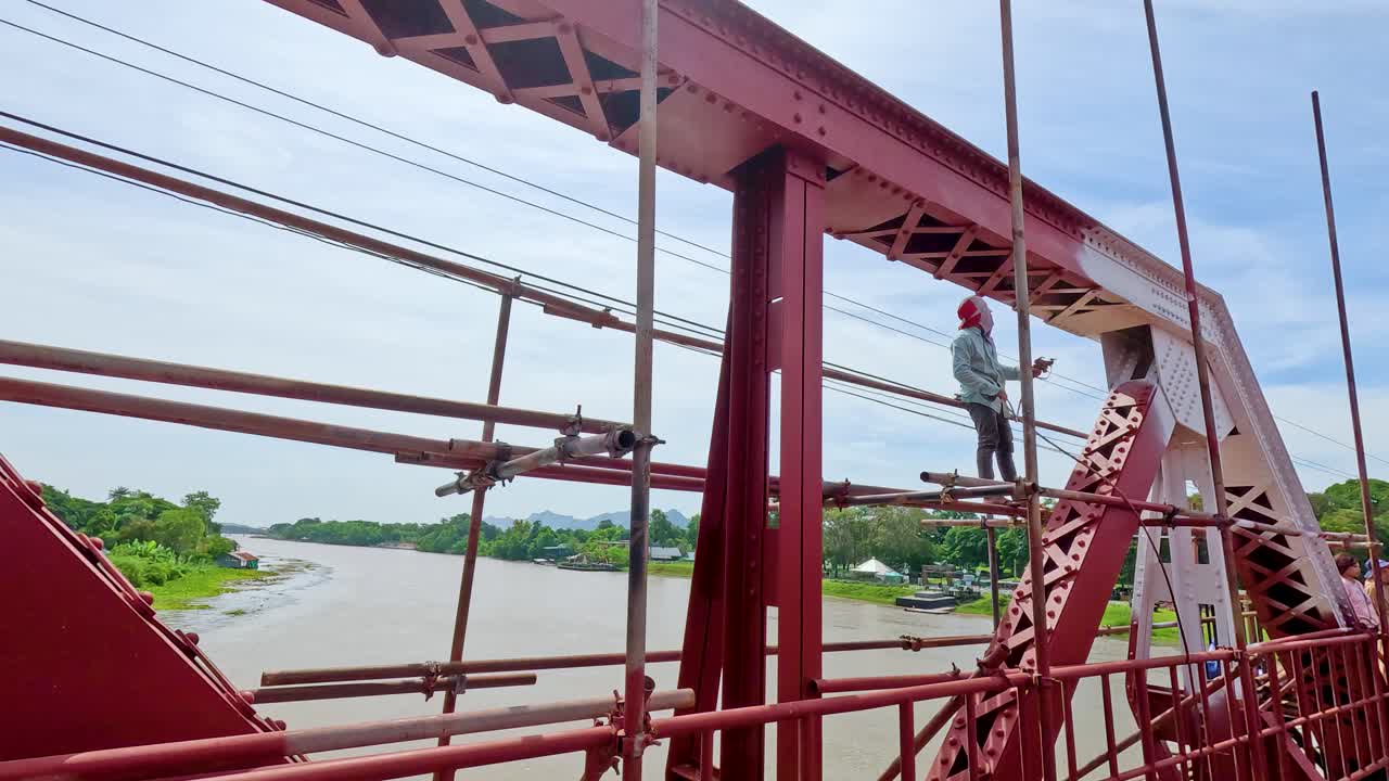 A worker stands on scaffolding, painting a red steel bridge above a wide river under bright daylight. Camera pans steadily, revealing scenic surroundings