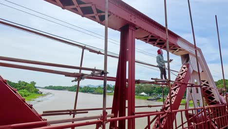 a worker stands on scaffolding, painting a red steel bridge above a wide river under bright daylight. camera pans steadily, revealing scenic surroundings