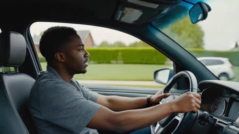 young adult african american male focused while driving sedan through quiet suburban neighborhood, looking straight ahead through windshield during daytime journey