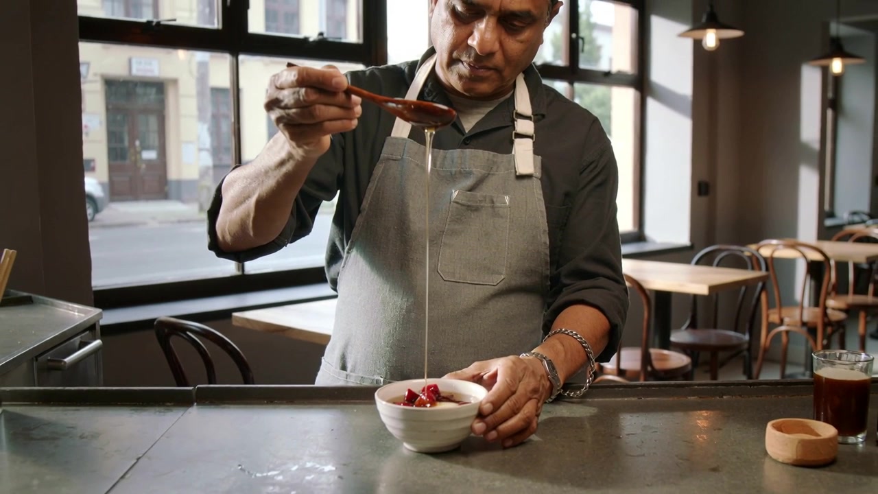 Uno chef con grembiule grigio, usando un cucchiaio, ricopre la frutta con sciroppo in una ciotola al bancone del caffè, preparando un piatto.