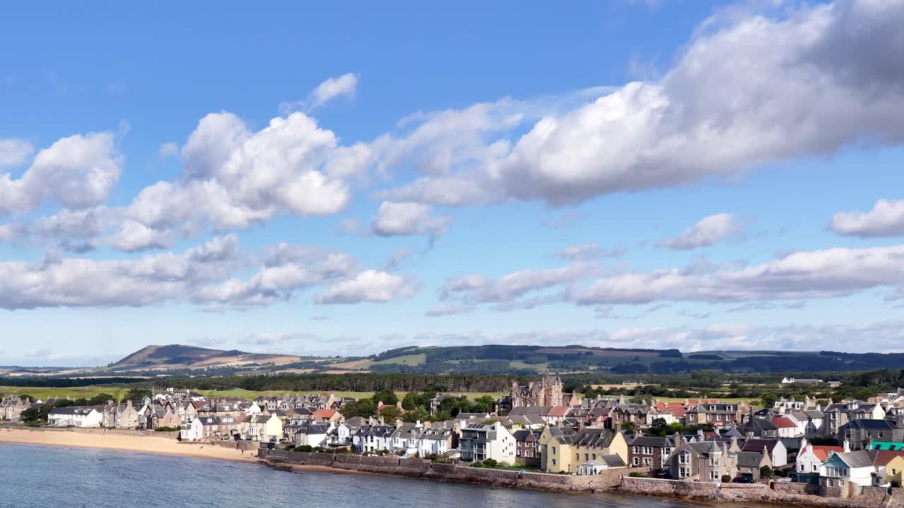 Drone camera smoothly pans across waterfront village, harbor, and beach under bright daylight skies