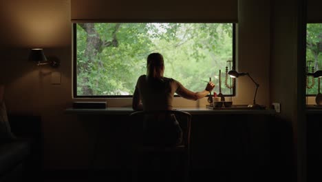 a woman picking up a pencil and writing on a notepad at a desk in front of a wide window with a natural green landscape outside