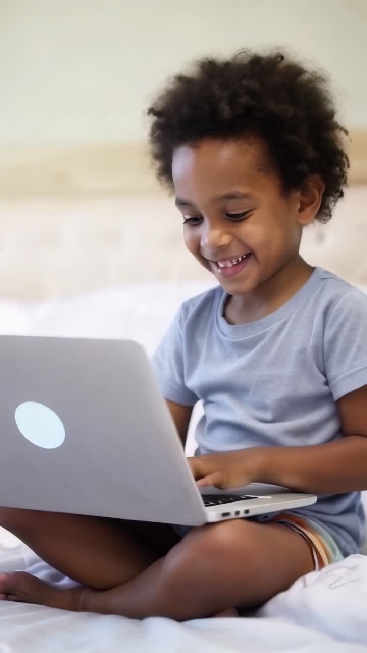 Smiling afro boy playing online with a laptop sitting on the bed.