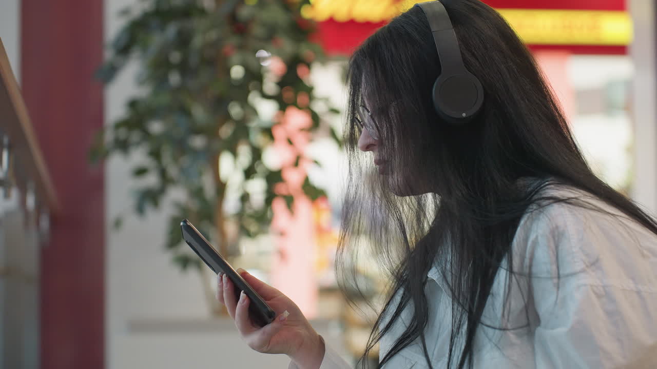 Side view of young lady in white shirt holding phone, gently dancing to music with headphones on, illuminated by blur of vibrant mall lights and indoor plant in background