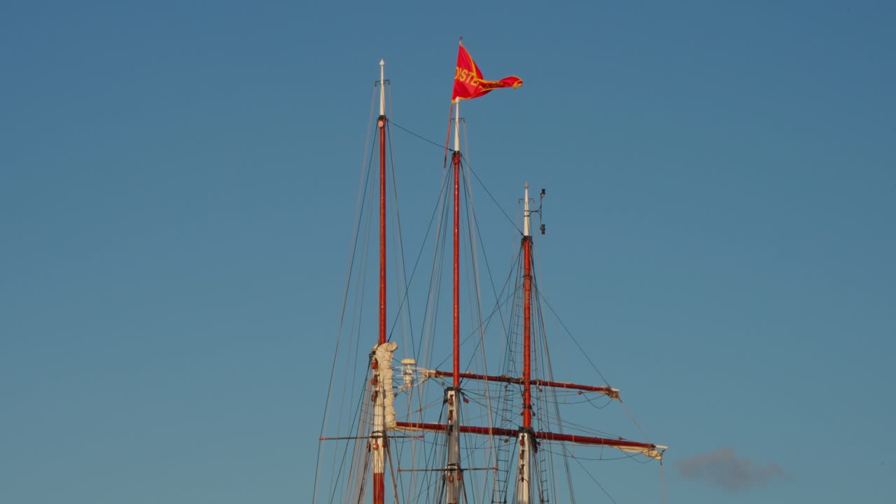Bright red flag with gold detail flying atop a tall wooden mast of a classic ship under clear blue sky. Location: Amsterdam, Netherlands (Nederland)