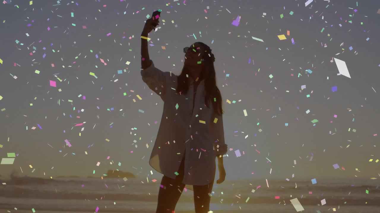 Donna che cammina sulla sabbia con coriandoli che fluttuano e solleva lo smartphone per un selfie sui social media al tramonto.