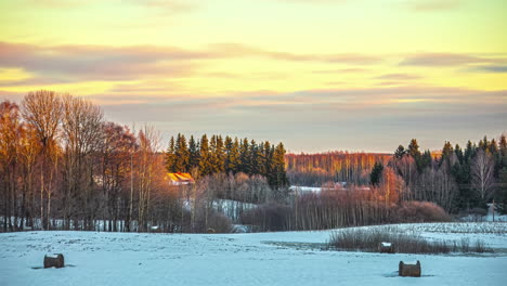 timelapse of a snowy landscape with trees and wooden cabins and with orange clouds advancing to the left in a golden sky