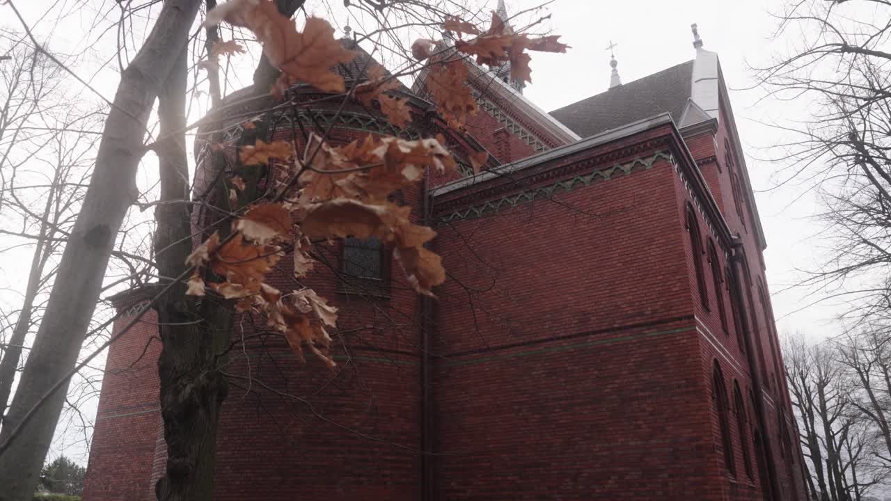 Large, red brick building with intricate architectural designs, surrounded by bare trees and fluttering leaves under an overcast sky, creating a somber atmosphere