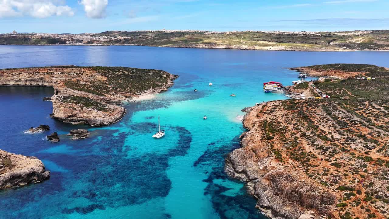 Vista aerea della Laguna Blu a Comino, Malta, che mostra acqua turchese, una costa rocciosa e diverse piccole imbarcazioni ancorate in una baia calma sotto un cielo sereno e soleggiato.