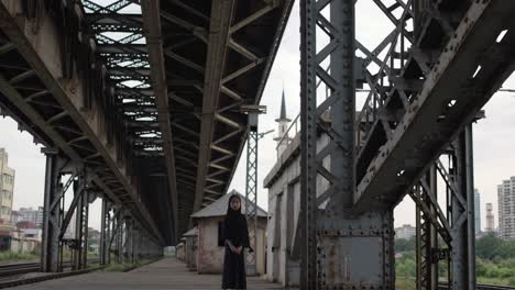 young girl wearing traditional clothing stands on a walkway beneath a large metal railway bridge, showcasing the contrast between traditional life and urban infrastructure