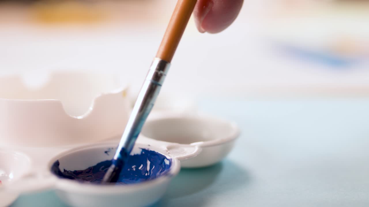Close-up of hand dipping brush in blue paint, soft lighting, shallow focus, creative workspace