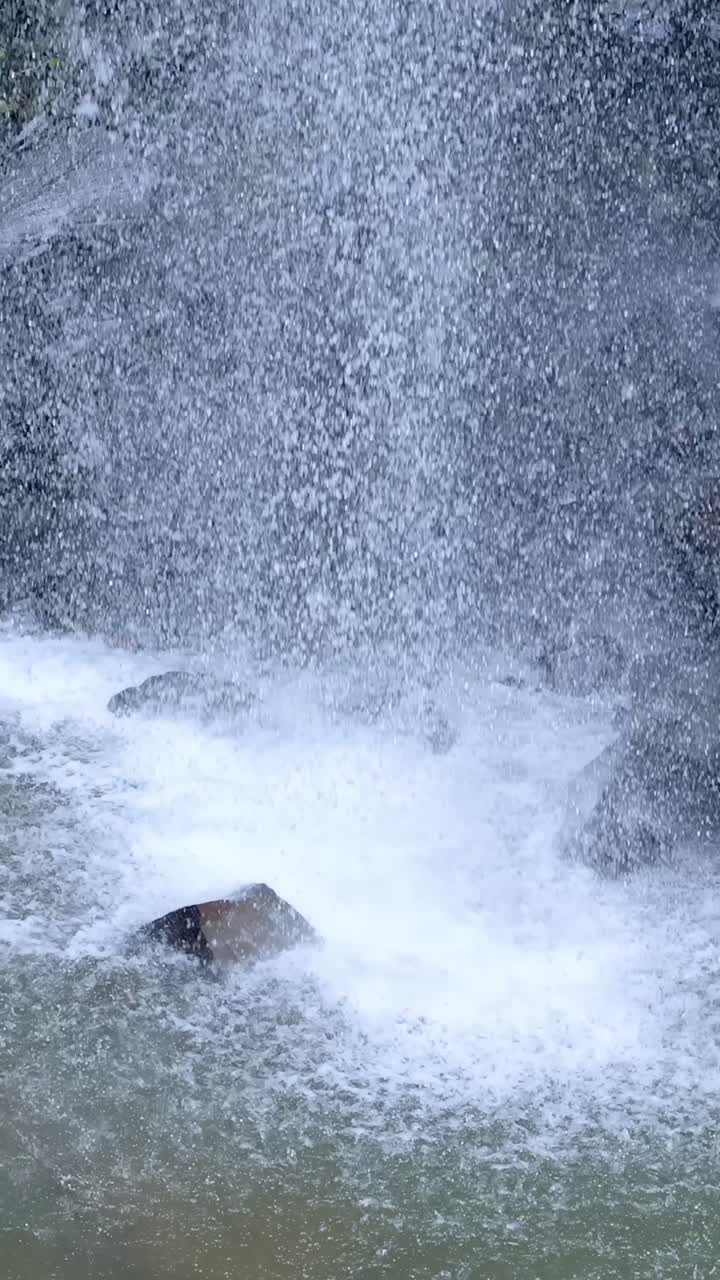 A steady camera captures a powerful waterfall crashing onto rocks in a lush forest setting, with bright daylight and dynamic water movement throughout