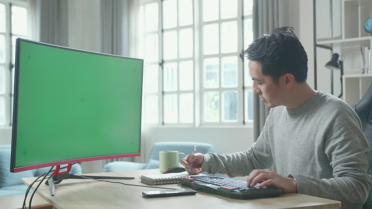 Asian Man In Long Sleeved T-Shirt Looking At Green Screen Desktop Computer And Writing In Notebook On A Table While Working At Home.