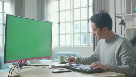 asian man in long sleeved t-shirt looking at green screen desktop computer and writing in notebook on a table while working at home.