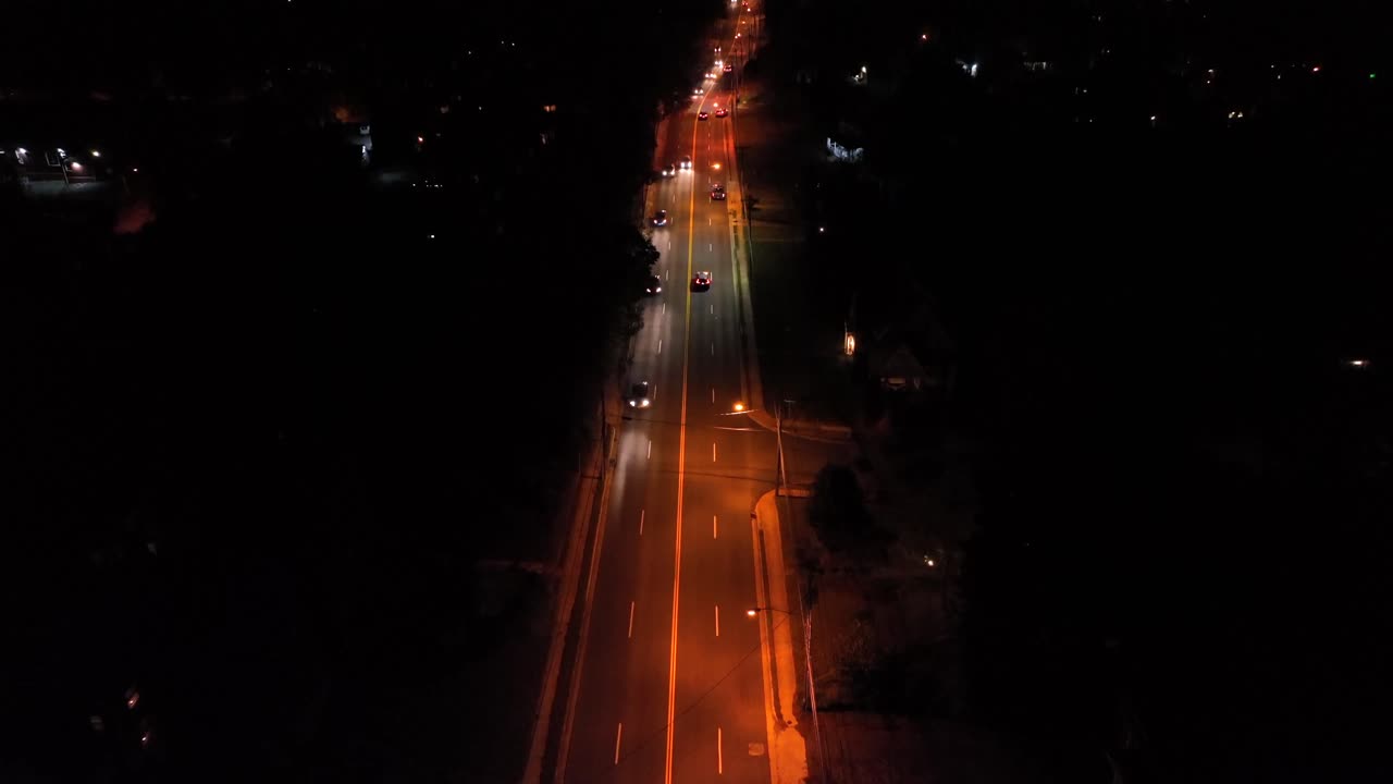 Residential America night scene with a long illuminated roadway, moving cars, orange streetlights and dark surrounding neighborhood area. drone wide shot. Interstate highway in usa at midnight