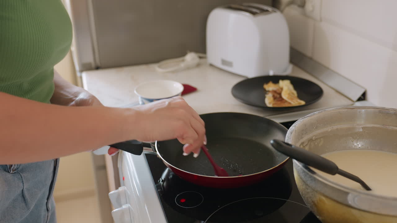 Close up of woman in green shirt preparing frying pan with red spatula beside bowl of batter and plate of cooked pancakes in bright kitchen atmosphere with toaster in background