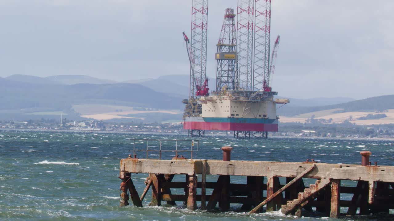 Static shot of oil platform beyond weathered pier, daylight, choppy sea, distant Scottish Highlands