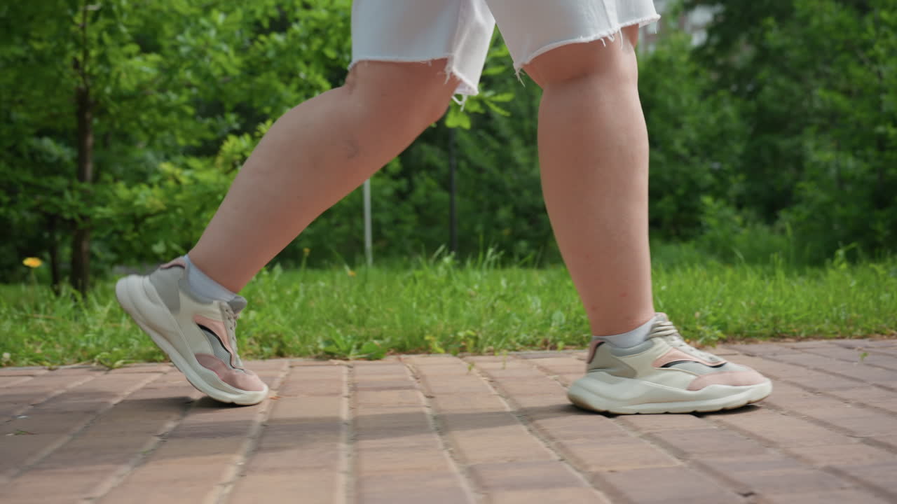 밝은 여름날, 무성한 녹색 나무와 잔디로 둘러싸인 벽돌길을 따라 운동화와 흰 반바지를 입고 평화로운 야외 산책을 즐기는 뚱뚱한 여성의 측면 모습