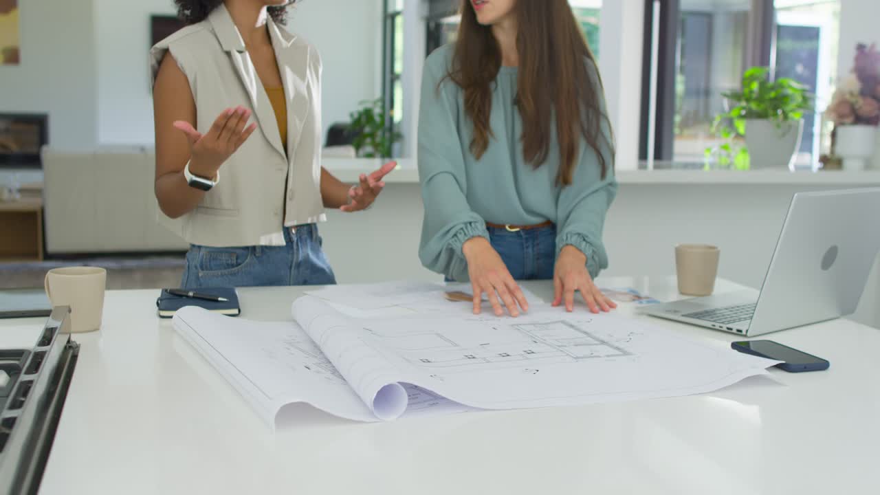 Diverse female colleagues unrolling plans, smoothing at white counter, using laptop for design