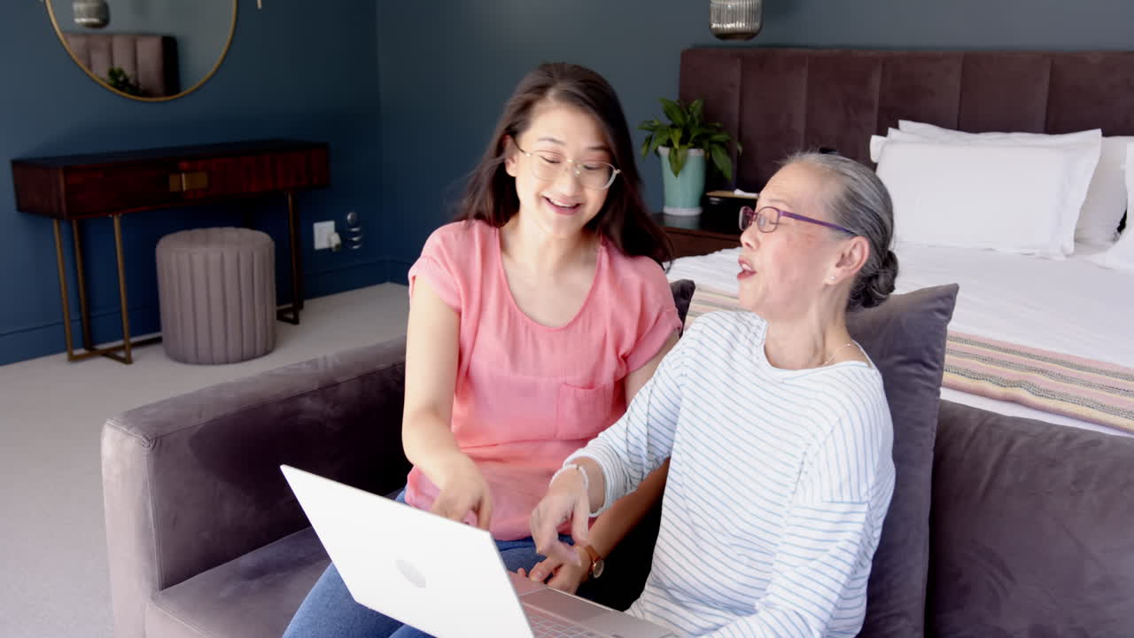 Using laptop, Asian grandmother and granddaughter smiling and working together in modern bedroom