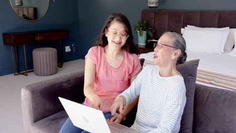 using laptop, asian grandmother and granddaughter smiling and working together in modern bedroom