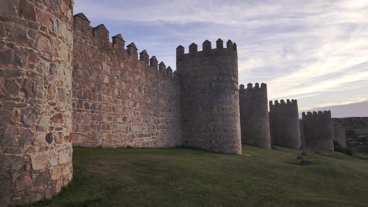 Ampia ripresa a mano delle iconiche mura del castello di Ávila in Spagna, bagnate dalla luce dorata del tramonto. Antiche torri di pietra e merlature si estendono su un paesaggio erboso, con un cielo drammatico sopra.