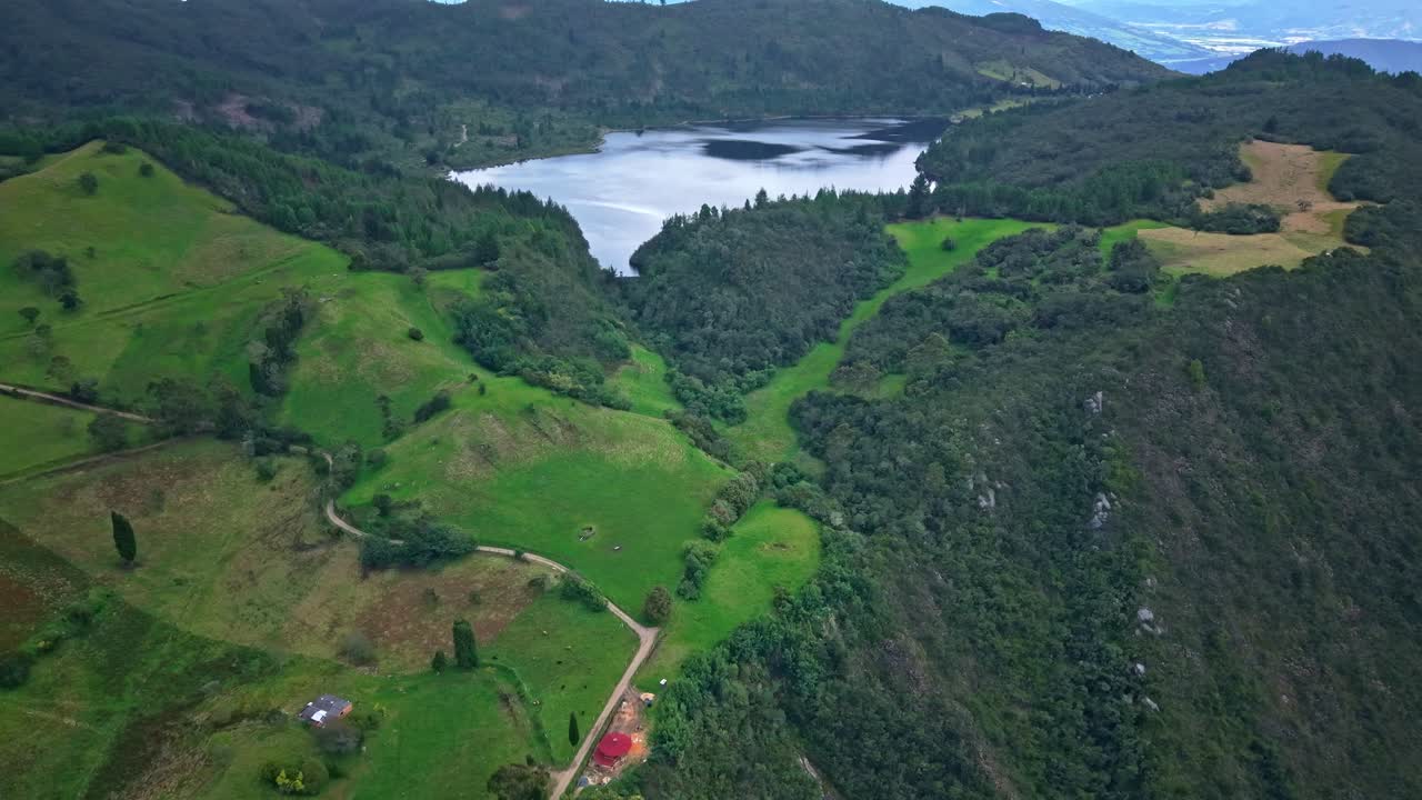 Drone pulls back from Laguna de Pantano Redondo, revealing forest edges, green fields and surrounding hills in Cundinamarca’s natural landscape