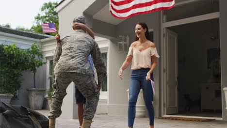 Happy-caucasian-male-soldier-lifting-son-and-greeting-wife-with-flag-hanging-outside-their-house