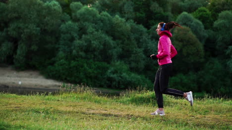 a girl in a pink jacket and black pants runs near the river in headphones preparing for the marathon