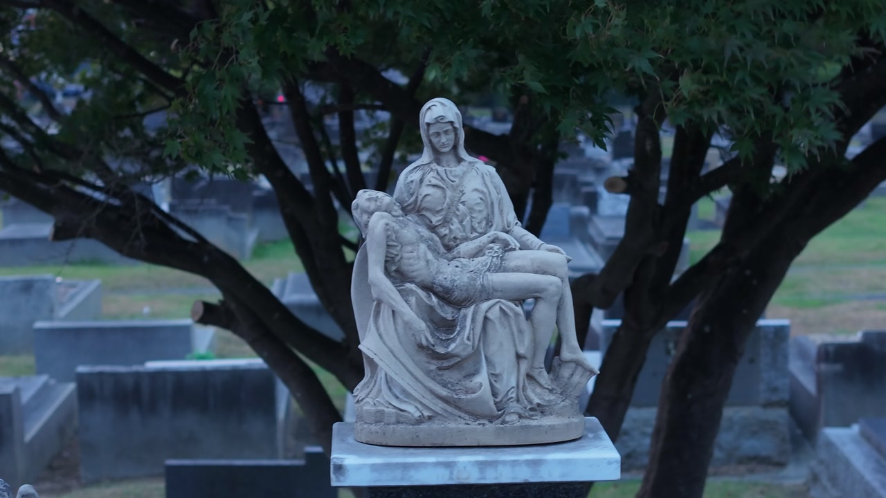 Tranquil cemetery scene in Melbourne during sunset, featuring solemn statues and a cross amid dusk lighting. Silence pervades the air, enhancing the atmosphere of reverence.