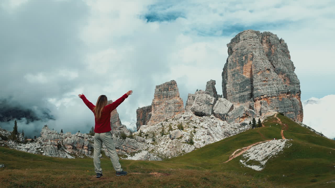 Victorious female hiker spreading arms wide, standing atop rocky mountain landscape with dramatic Dolomites peaks surrounding scenic alpine valley