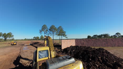 aerial fpv view of excavator digging soil trench beside concrete wall and pine trees