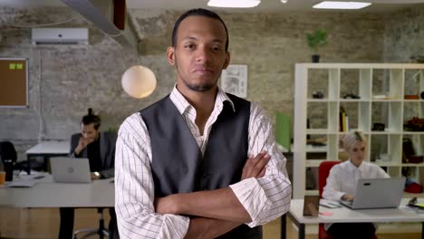 young african businessman is standing with arms crossed and watching at camera in office, colleagues are working with laptops, work concept