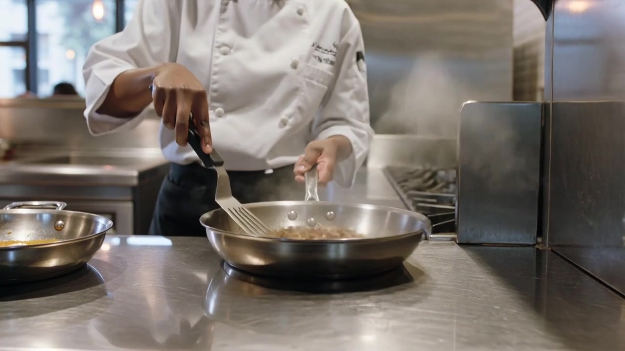 Chef in giacca che posiziona la padella sul fornello a gas, stabilizzando e girando il cibo con una spatola per finire di saltare
