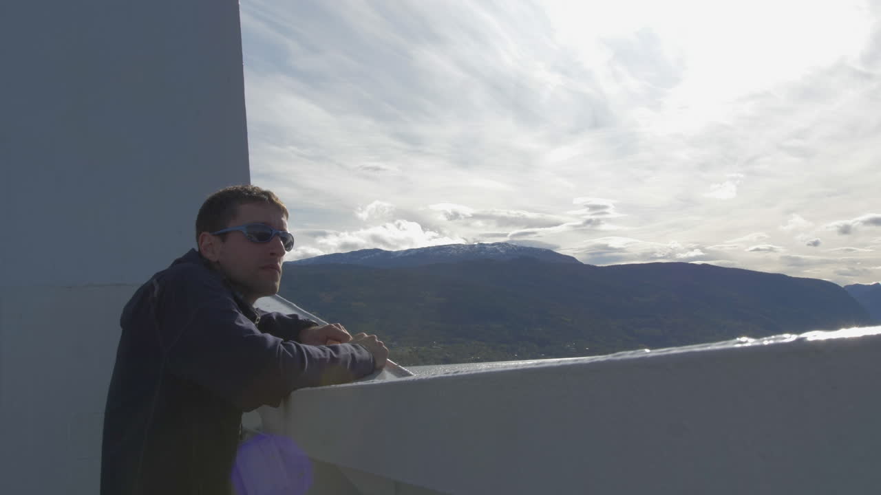 un hombre con gafas de sol mira al borde de un ferry con una bonita vista de las montañas en un día soleado, a cámara lenta