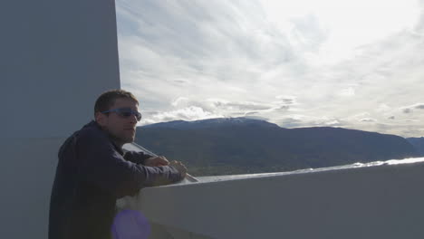 a man in sunglasses looks out over the edge of a ferry taking in a nice view of mountains on a sunny day, slow motion