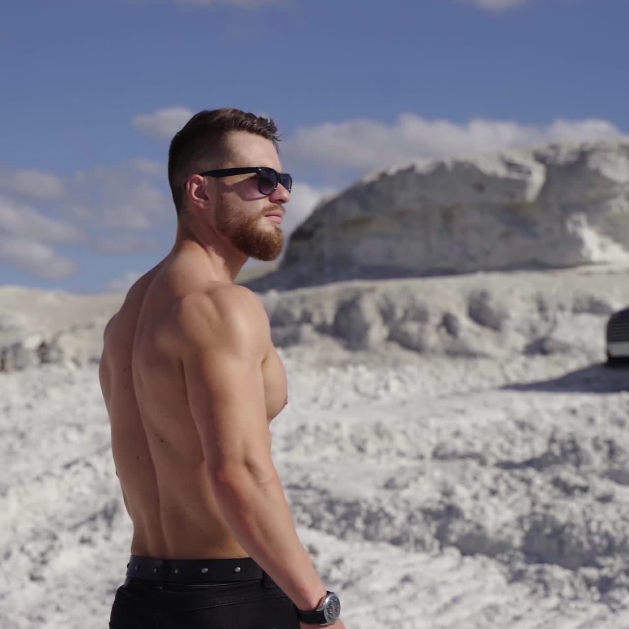 Muscular sportsman in the mountains. Handsome shirtless athlete in sunglasses on a white canyon on the background of luxury black car in sunny day.