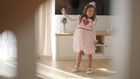 a young girl dances joyfully in a bright, cozy living room. she wears a pink dress with a heart design, moving playfully in the sunlit, minimalist space with neutral decor and soft natural light.