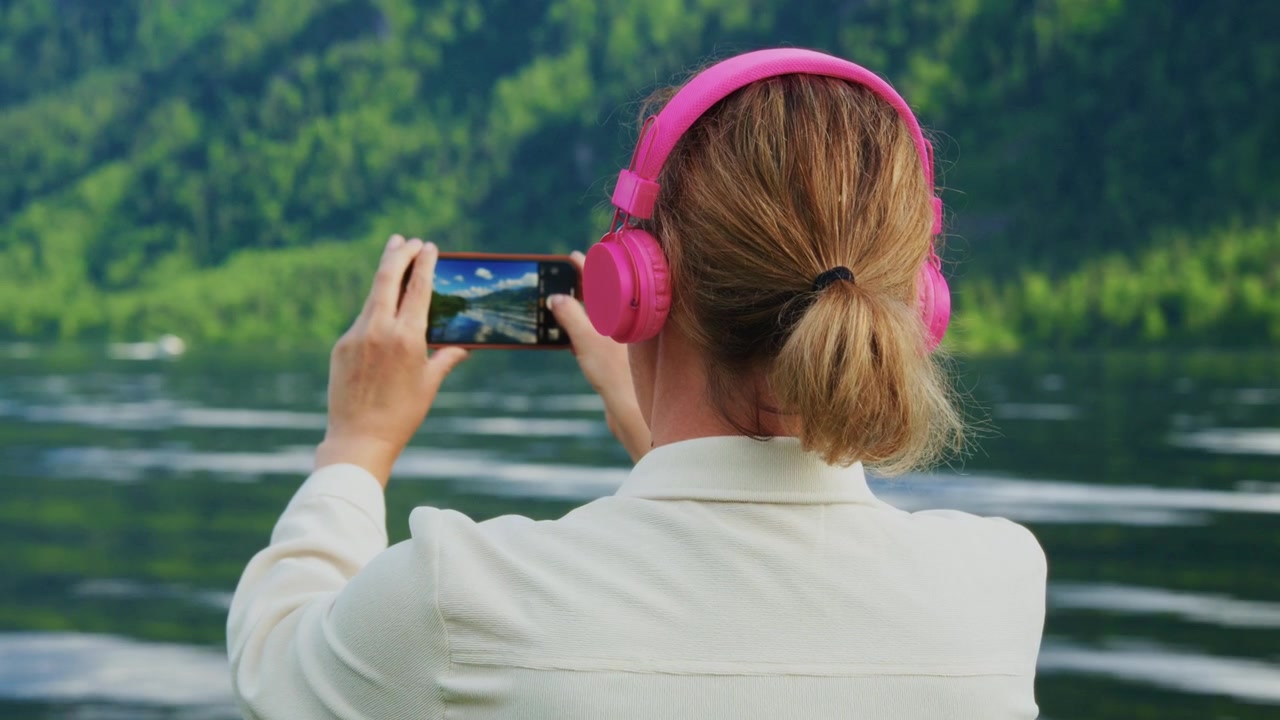 Woman wears pink headphones, takes photos by lake. She stands calm, focused on capturing scenic moments. Vibrant green mountains surround tranquil water surface. Ideal for travel, relaxation