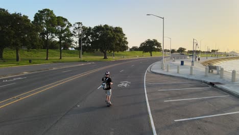 young man strolling at empty lakeshore drive with electronic skateboard at dusk. - aerial, rear